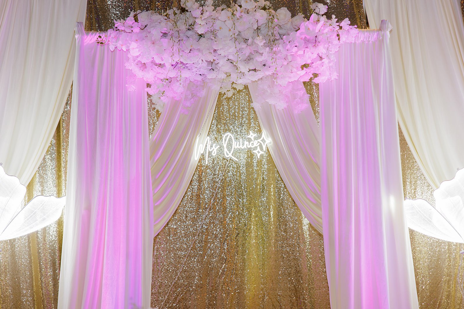 Arco floral con cortinas blancas, lentejuelas doradas y un letrero de neón en una quinceañera en Admiral Theater, Omaha