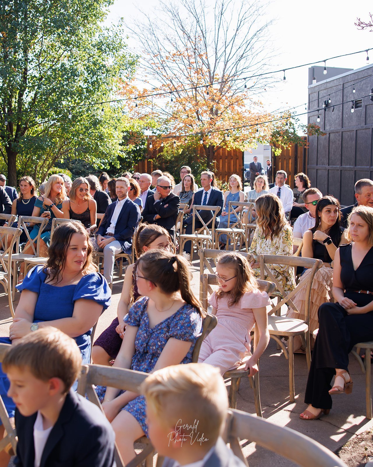 Wedding guests seated in wooden cross back chairs under string lights at an outdoor ceremony at The Arbor Event Venue, Omaha