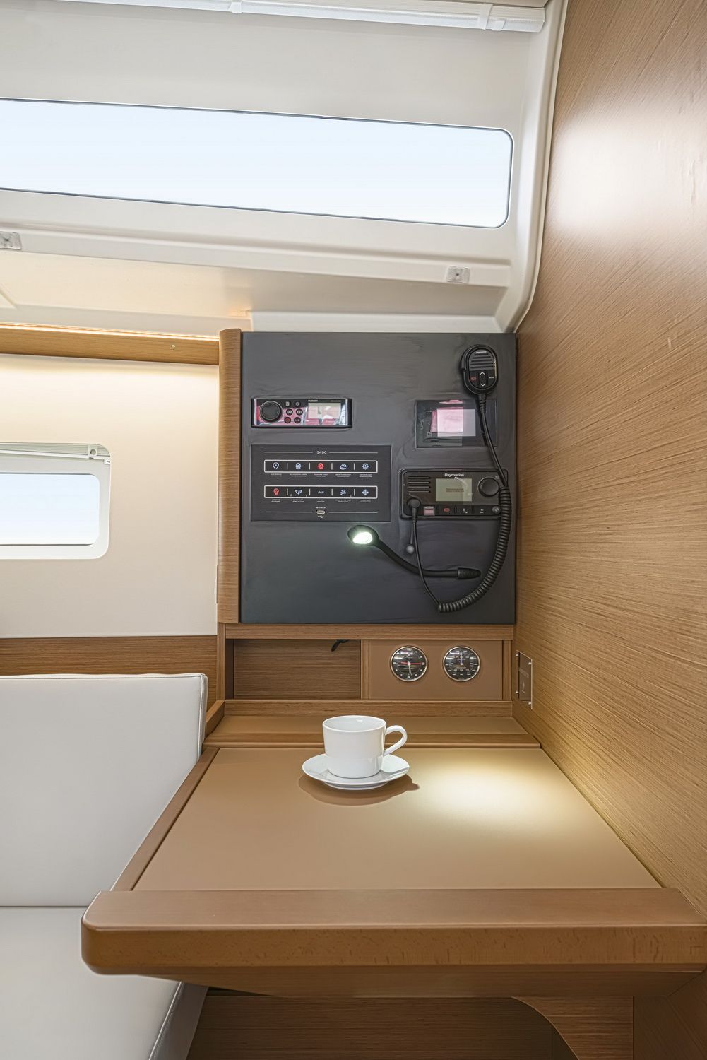 Panel nawigacyjny z elektroniką i radiem VHF, obok drewniany stół z filiżanką kawy, w jasnym wnętrzu jachtu.