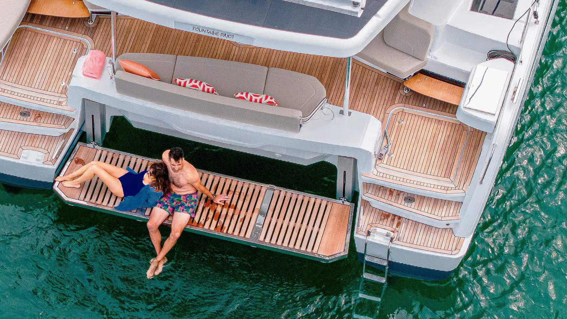 Ein Paar entspannt auf der ausklappbaren Heckliege des Katamarans Fountaine Pajot FP41, die im Wasser liegt.