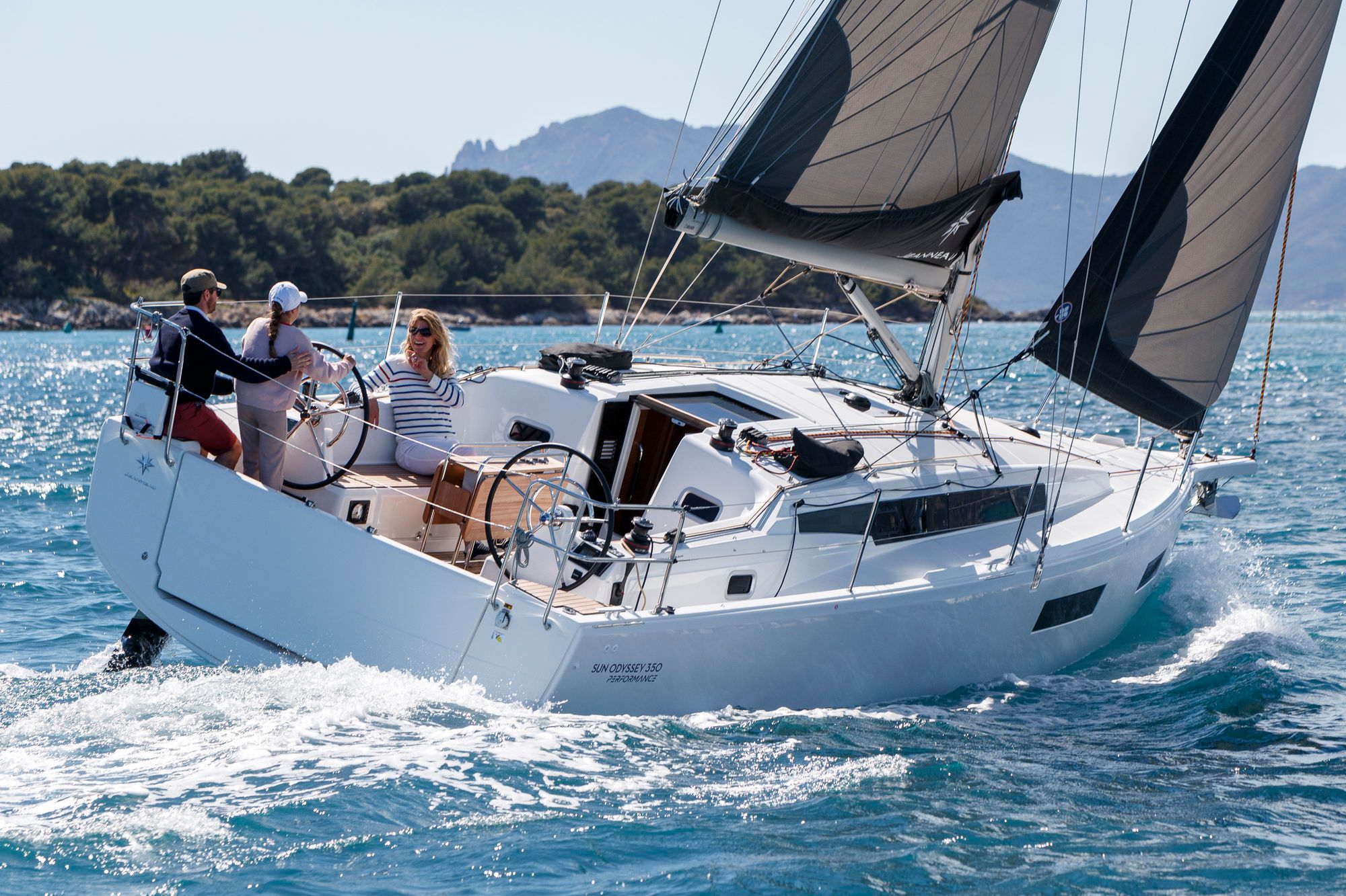 Jacht żaglowy Sun Odyssey 350 pod ciemnymi żaglami - załoga w kokpicie z dwoma kołami sterowymi, w tle skaliste wybrzeże.