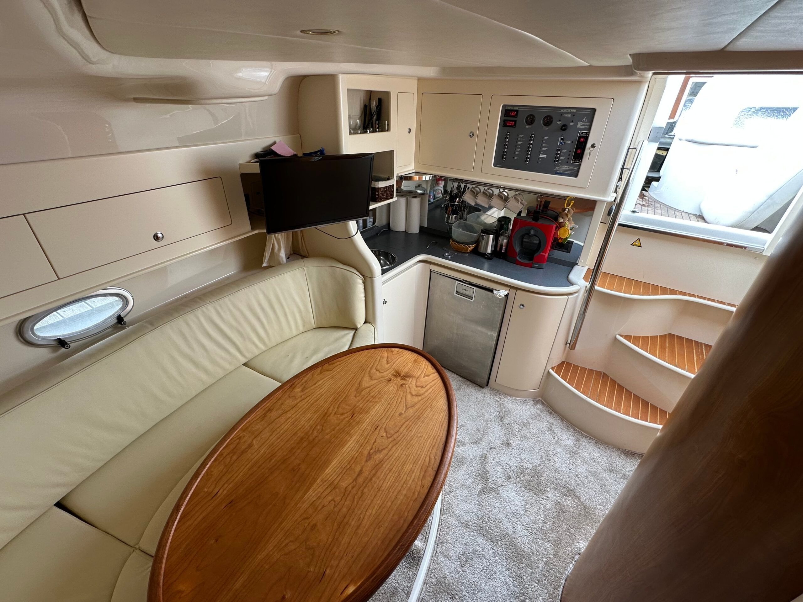 Widok na mesę i kambuz jachtu Sessa 35 Oyster. Widoczne beżowe kanapy, drewniany stół, telewizor, lodówka i panel elektryczny.