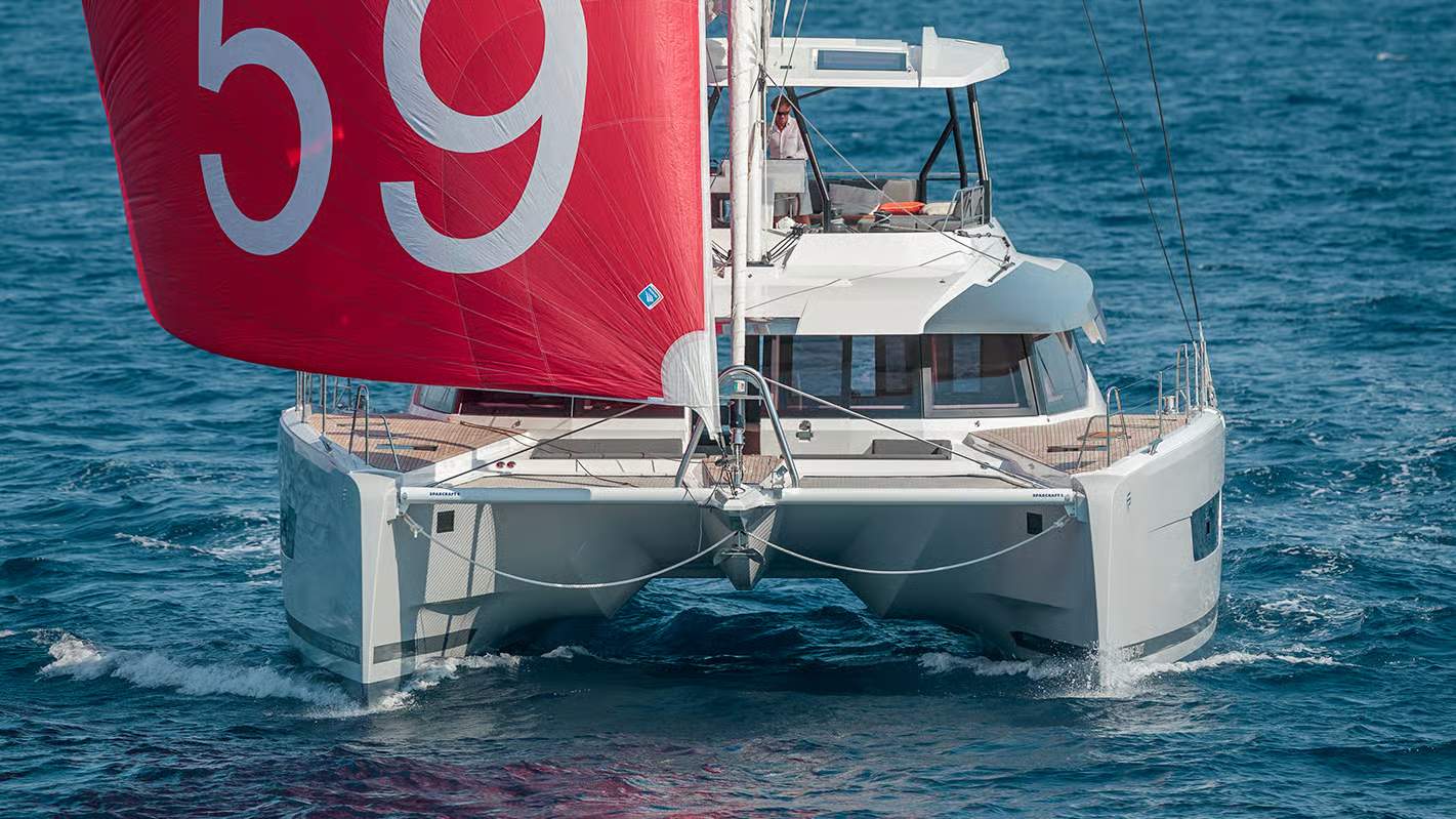 Fountaine Pajot Samana 59, katamaran żaglowy płynący po morzu z rozłożonym czerwonym żaglem. Widoczny dziób i flybridge z osobą.