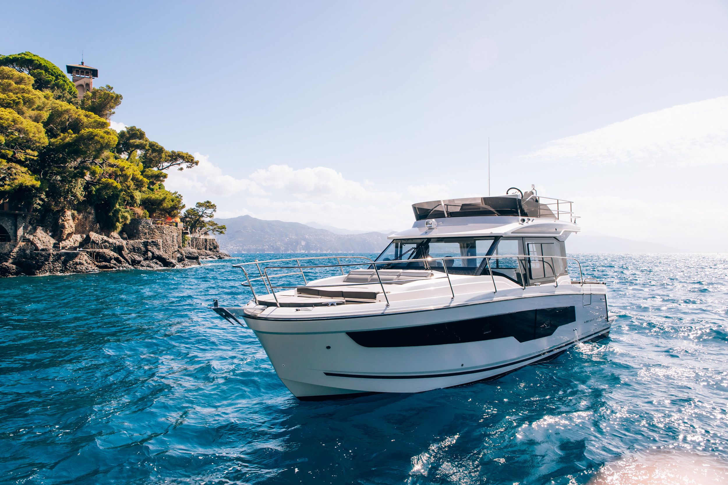 Jeanneau Merry Fisher 1095 Fly Serie2 motor yacht on blue water, front and side view, with the flybridge visible and a rocky coastline in the background.