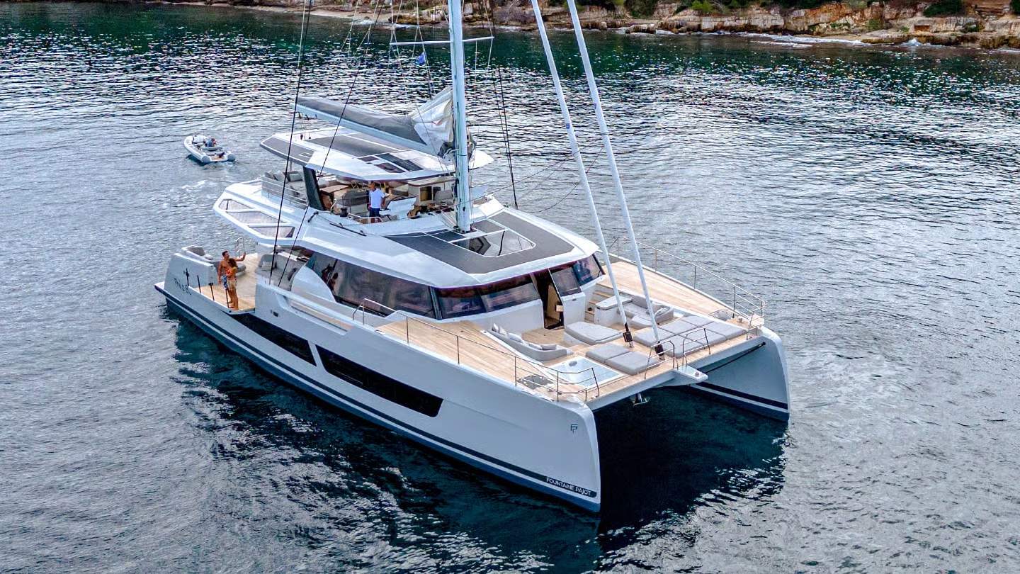 Duży biały katamaran Fountaine Pajot Thíra 80 na spokojnej wodzie, z otwartym bocznym balkonem i ludźmi na pokładzie, w tle skaliste wybrzeże.