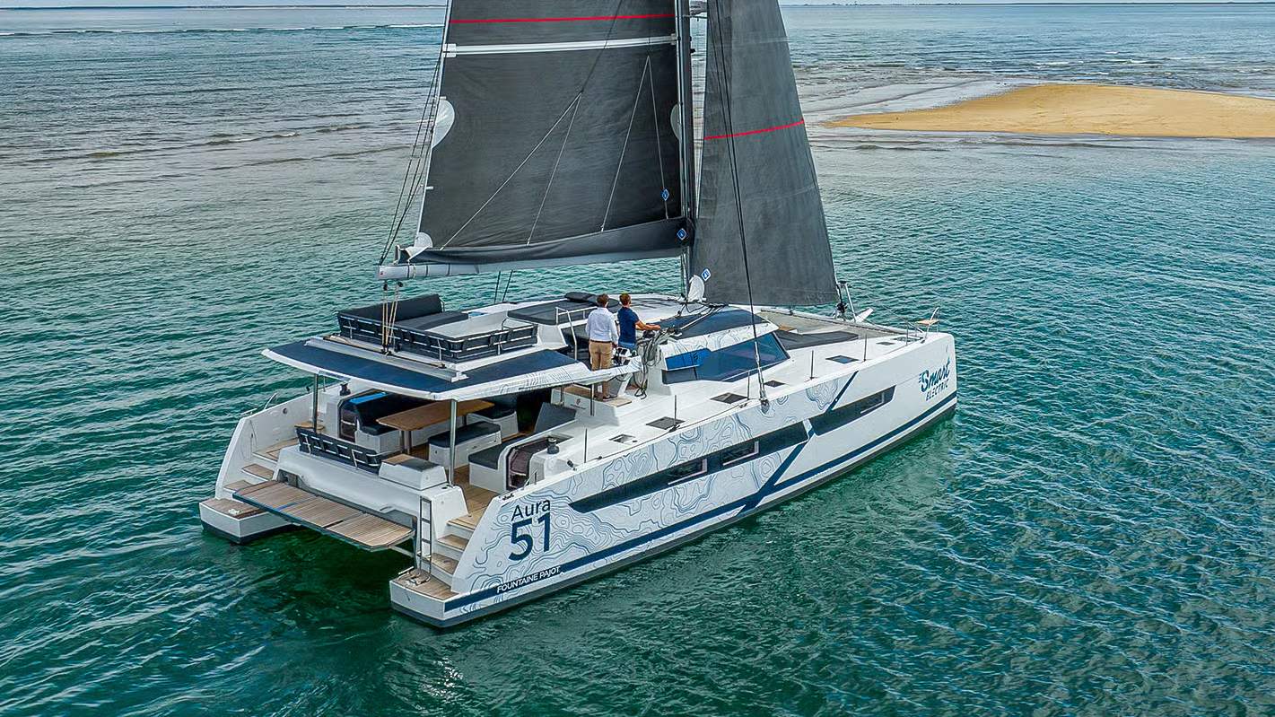 Katamaran Fountaine Pajot Aura 51 Smart Electric na wodzie, z dwoma osobami przy sterze i rozwiniętymi żaglami.