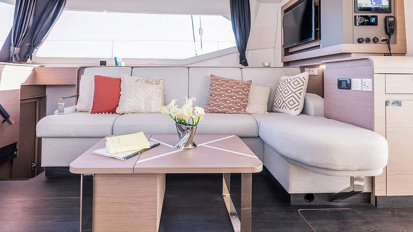Wnętrze salonu katamaranu Fountaine Pajot Aura 51 z dużą sofą w kształcie litery L, stolikiem kawowym i telewizorem.