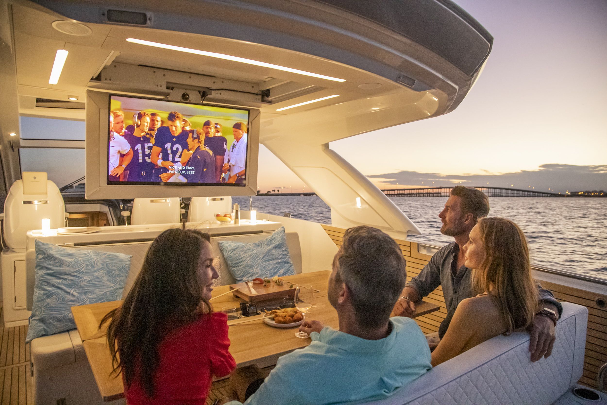 Four people relaxing in the cockpit of the Jeanneau DB/43 OB at sunset, watching TV. A bridge visible in the background.