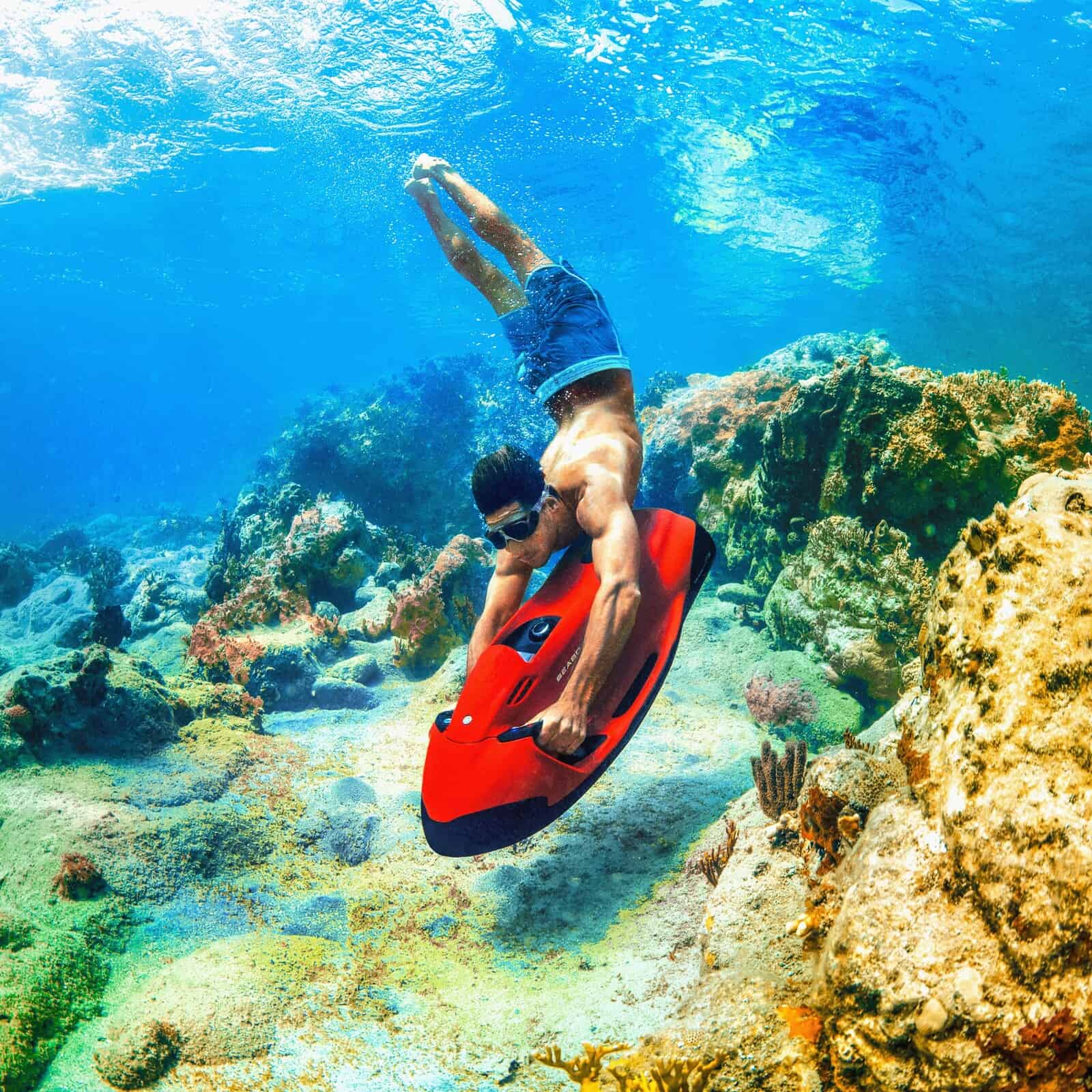 Ein Mann mit Schwimmbrille taucht mit einem roten Seabob F5SR im klaren, blauen Wasser über einem Korallenriff.