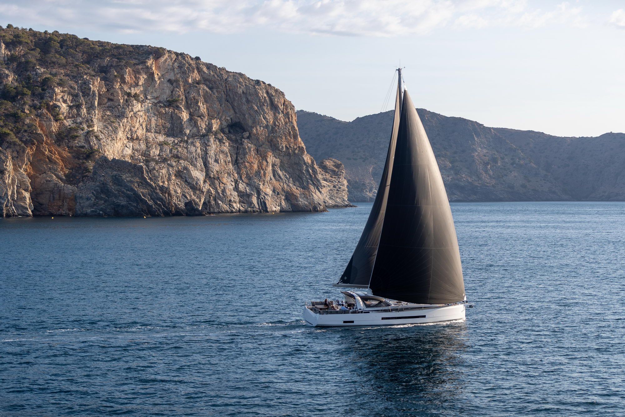 Biały jacht żaglowy Jeanneau Yachts 55 z czarnymi żaglami płynący po morzu wzdłuż skalistego wybrzeża.