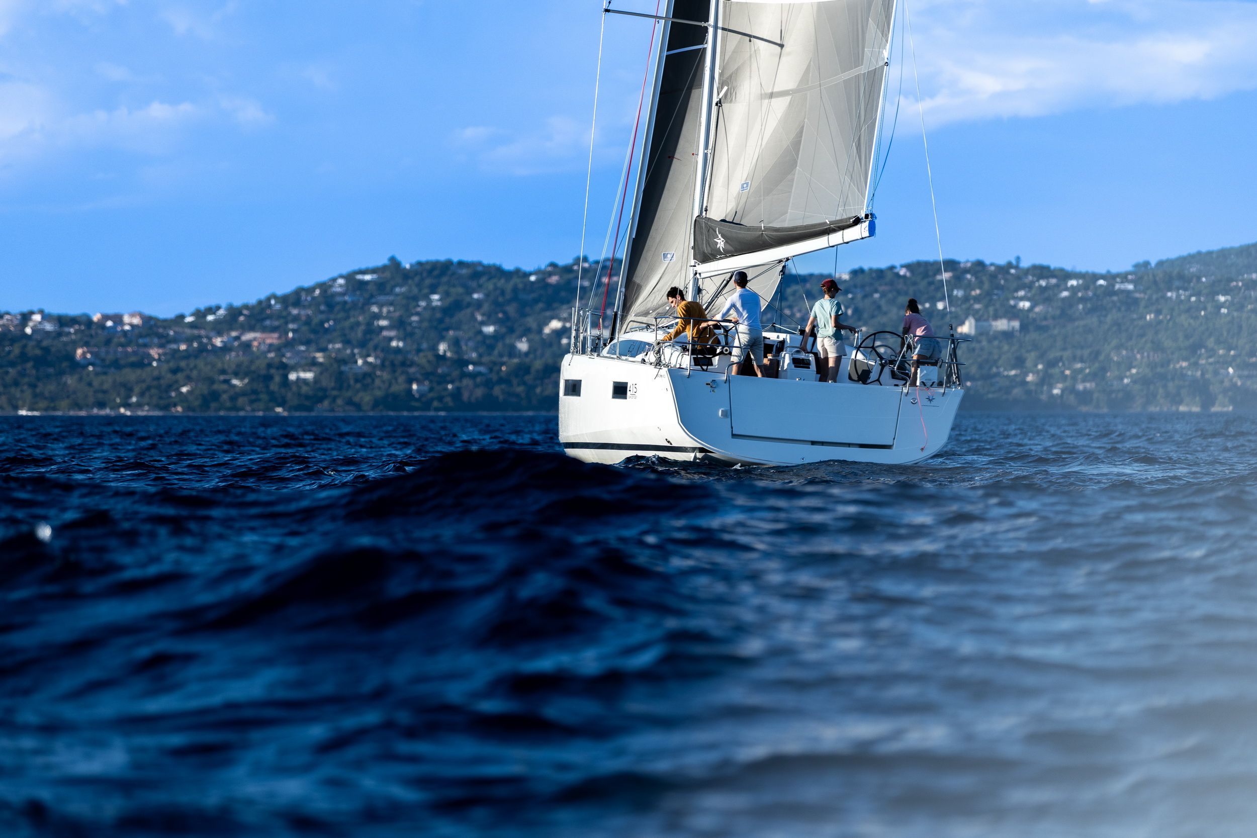 Jacht żaglowy Jeanneau Sun Odyssey 415 z załogą na otwartej wodzie, z rozwiniętymi żaglami, na tle górzystego wybrzeża.