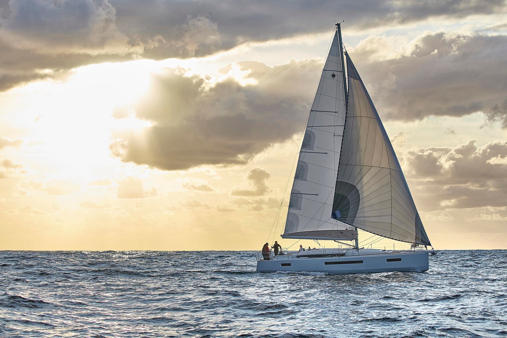 Jacht żaglowy Jeanneau Sun Odyssey 490 płynący po morzu o zachodzie słońca, z załogą na pokładzie.