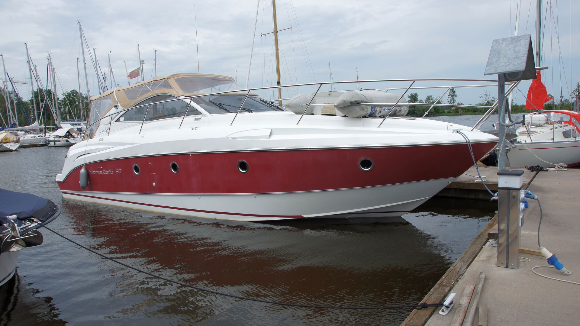 Beneteau Monte Carlo 37 Open zacumowany przy drewnianej kei, widok z burty. Jacht ma czerwony pas na kadłubie i beżowe cabrio.
