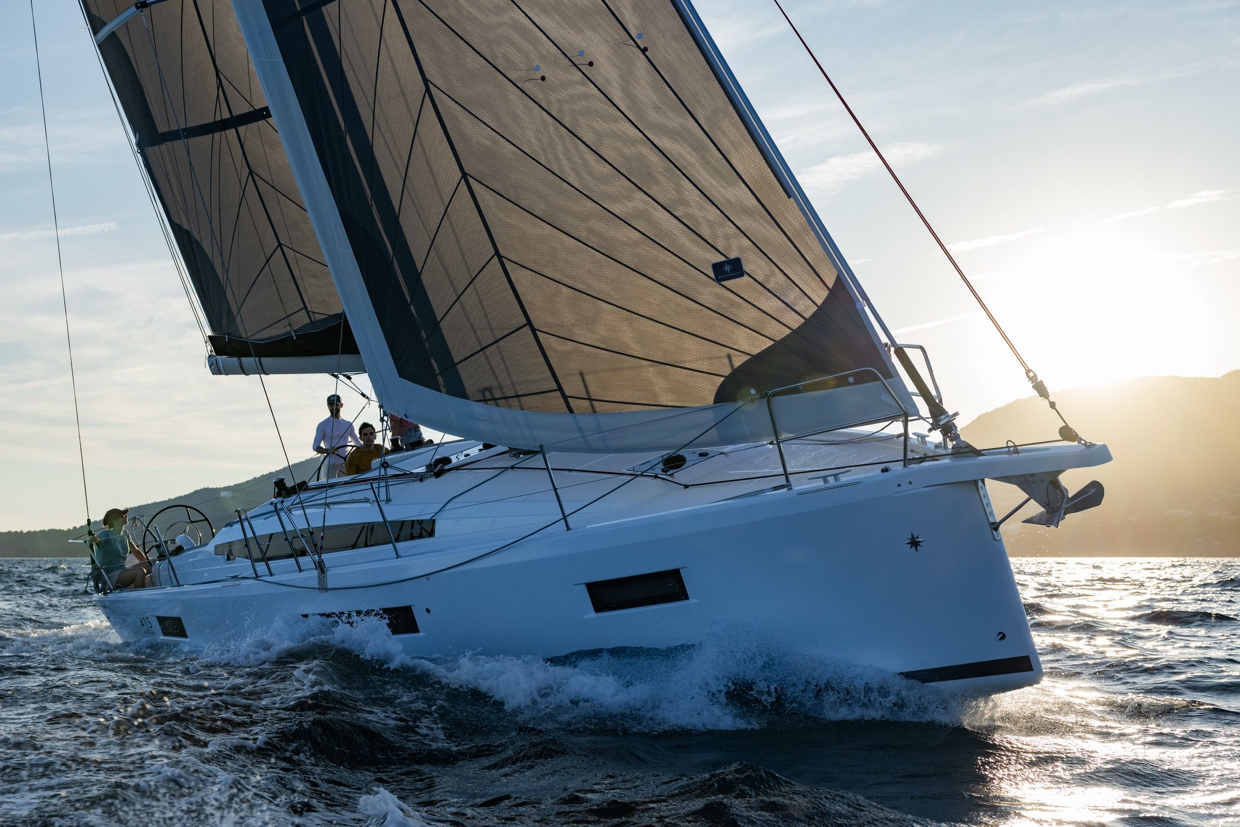 Jacht żaglowy Jeanneau Sun Odyssey 415 płynący po wodzie z podniesionymi żaglami, z załogą na pokładzie, o zachodzie słońca.