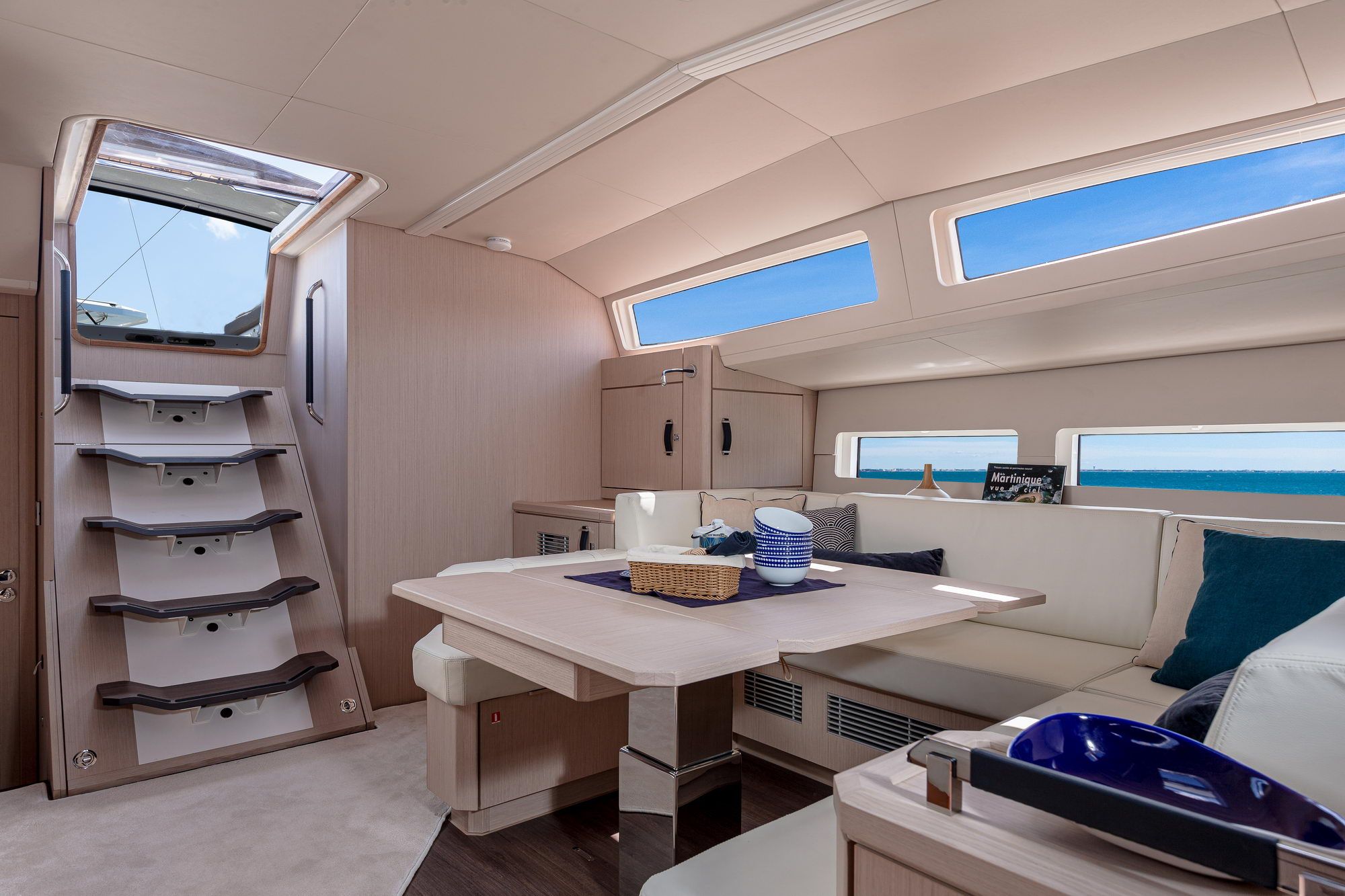 Wnętrze salonu jachtu Jeanneau Yachts 60 z jasnym stołem, kanapami, schodami i widokiem na morze przez okna.