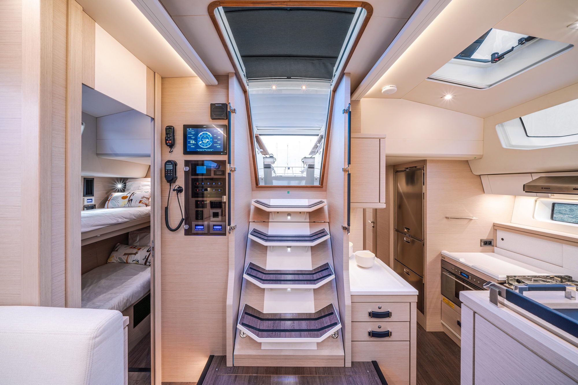 Wnętrze jachtu Jeanneau Yachts 65 z widokiem na schody, kabinę z łóżkami piętrowymi i kambuz. Widoczny panel sterowania.