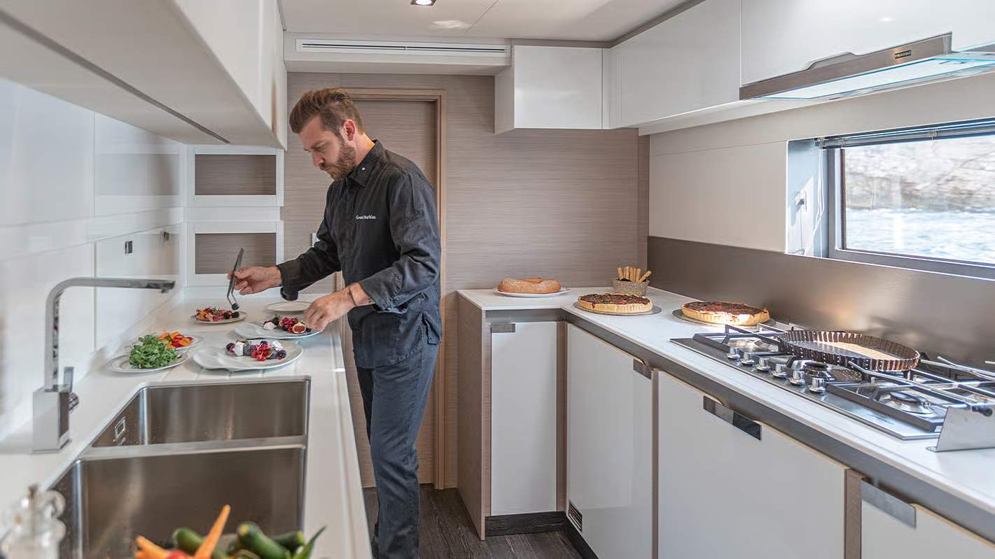 Mężczyzna przygotowujący posiłek w nowoczesnym kambuzie katamaranu Fountaine Pajot Samana 59, z widokiem na wodę.