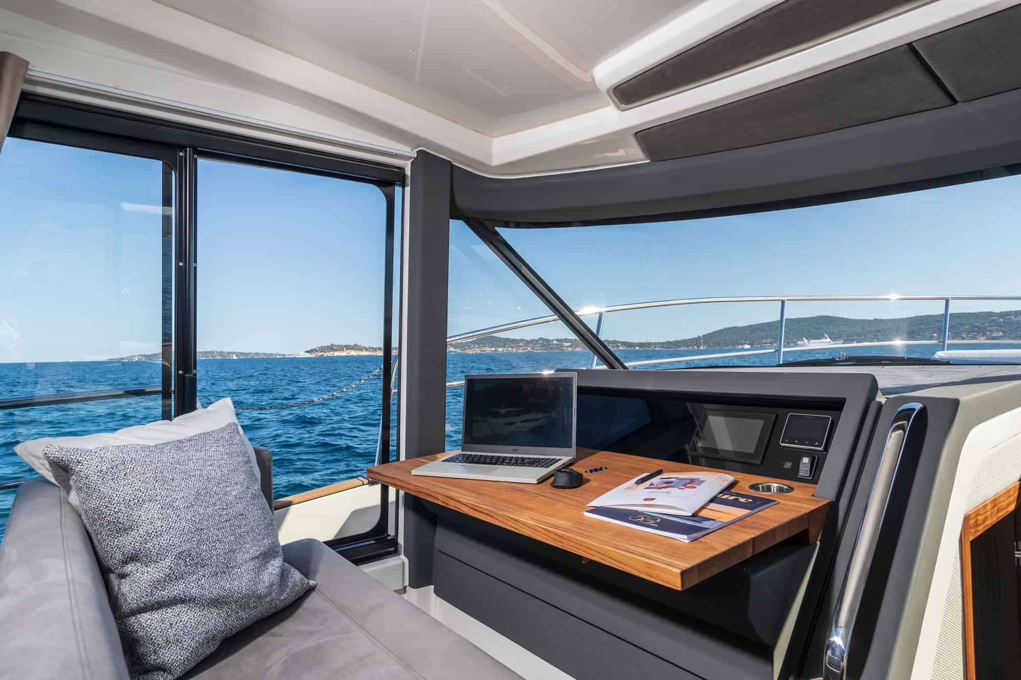 Interior of the saloon on the Marex 440 Gourmet Cruiser with a sea view. A small table with a laptop and a sofa are visible.