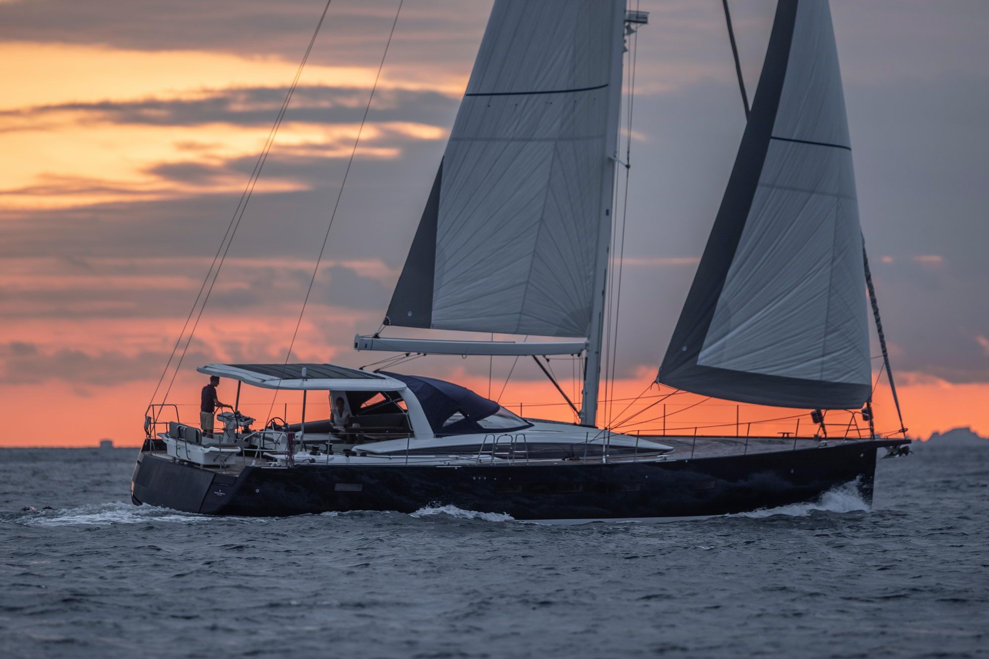 Jeanneau Yachts 65 płynący po morzu o zachodzie słońca, z rozwiniętymi żaglami i osobą za sterem.