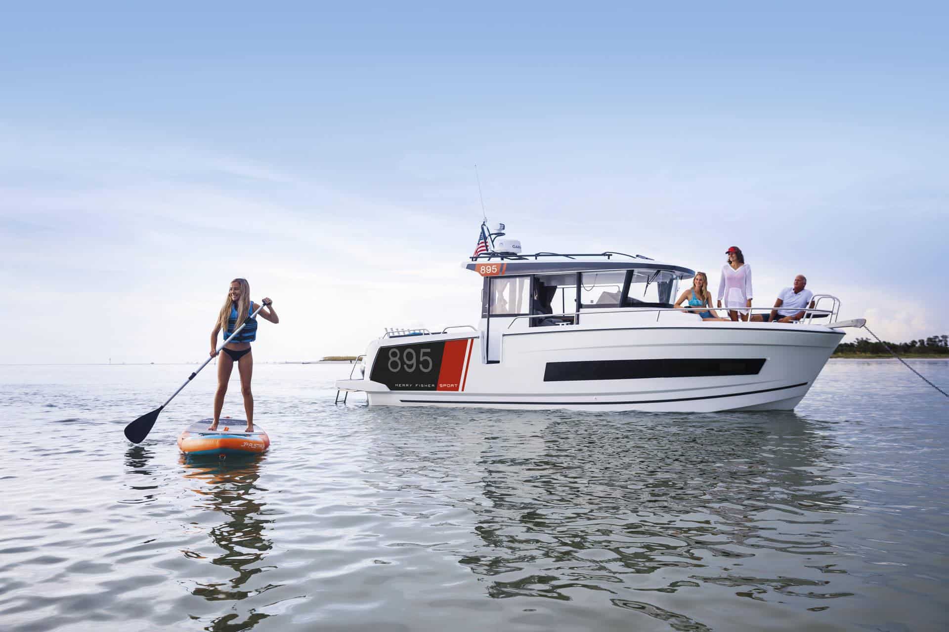 White Jeanneau Merry Fisher 895 Sport S2 motor yacht on calm water, with people on board and a girl on a SUP board alongside.
