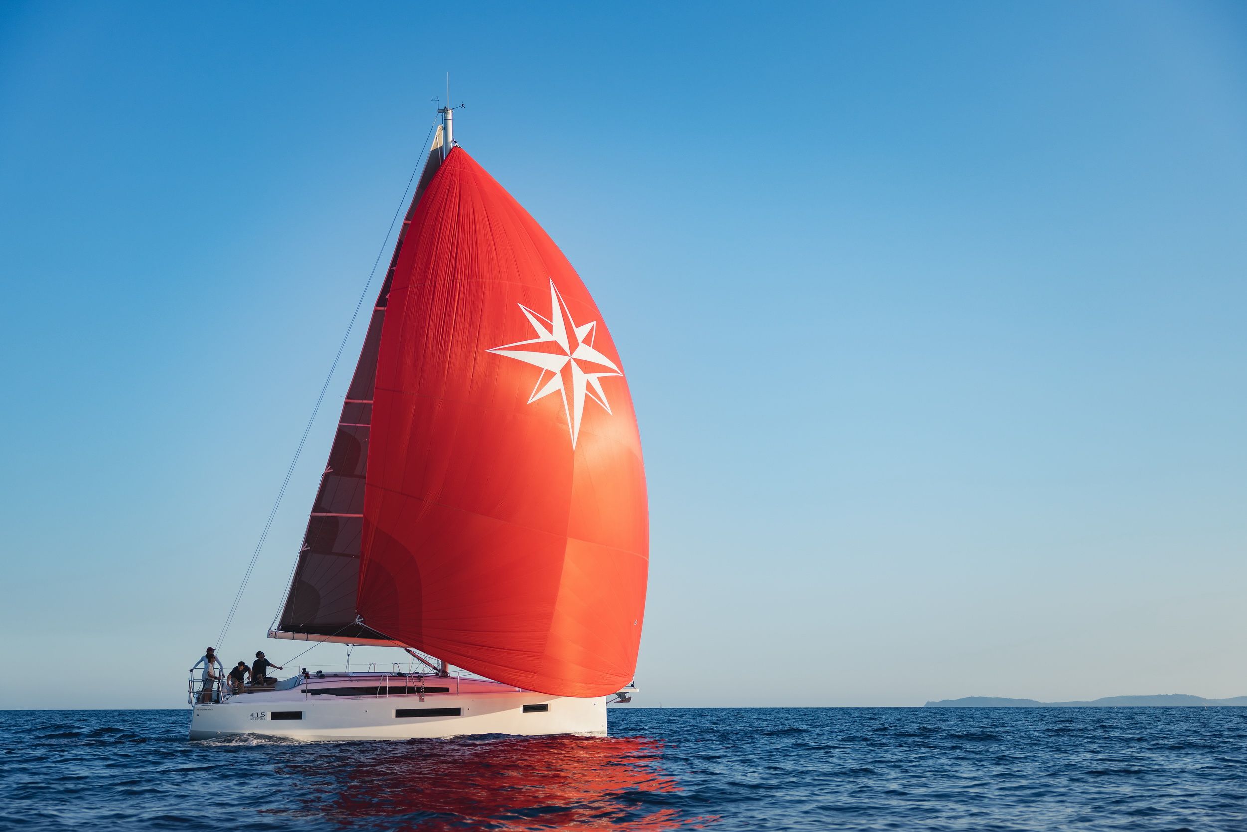 Jacht żaglowy Jeanneau Sun Odyssey 415 płynący po morzu z rozłożonym czerwonym spinakerem. Na pokładzie widać kilka osób.