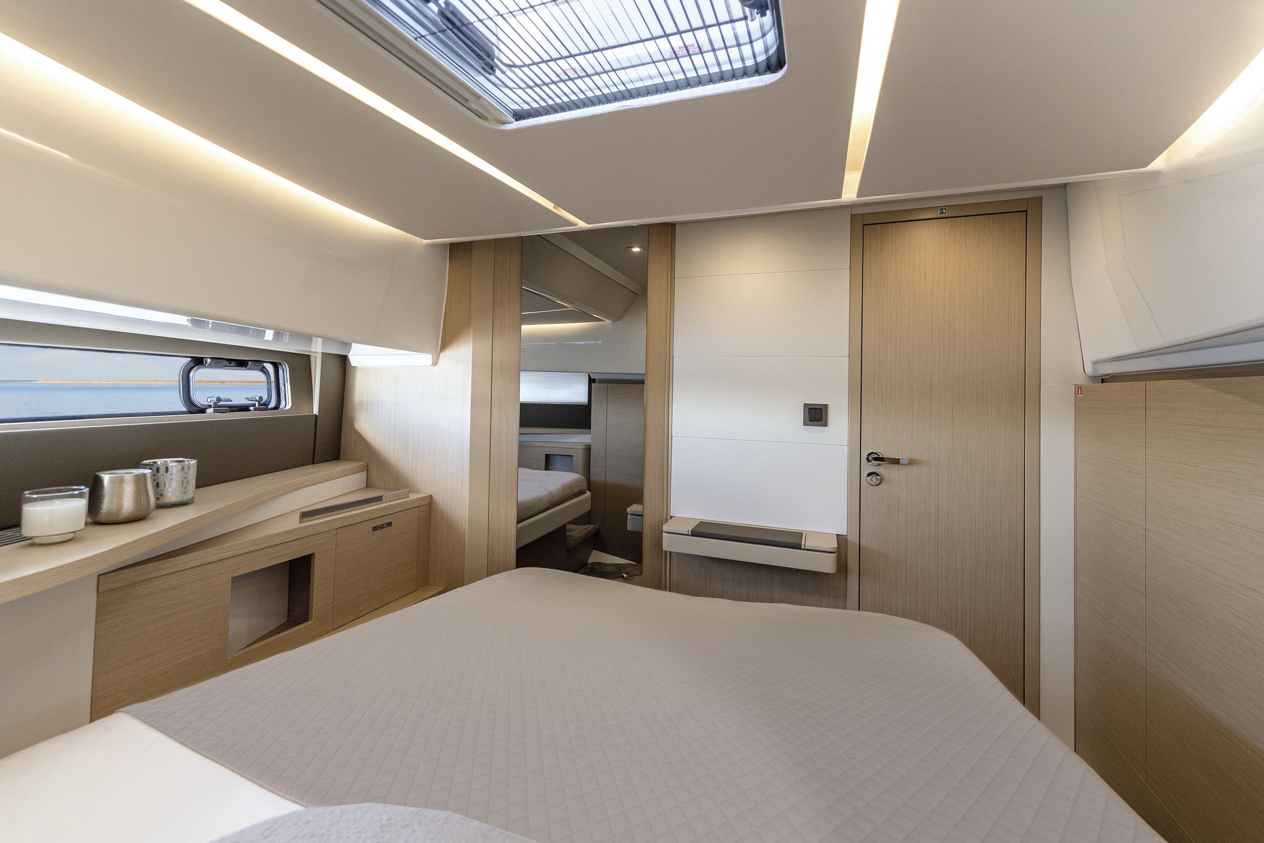 Innenansicht einer Schlafkabine der Jeanneau DB/37 IB mit Bett, Schränken, Fenster und Tür. Zu sehen sind moderne Beleuchtung und eine Dachluke.