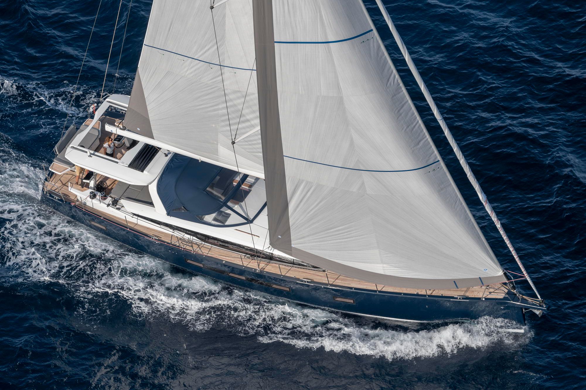 Widok z lotu ptaka na jacht żaglowy Jeanneau Yachts 65 z ciemnoniebieskim kadłubem, płynący po morzu z rozłożonymi żaglami.
