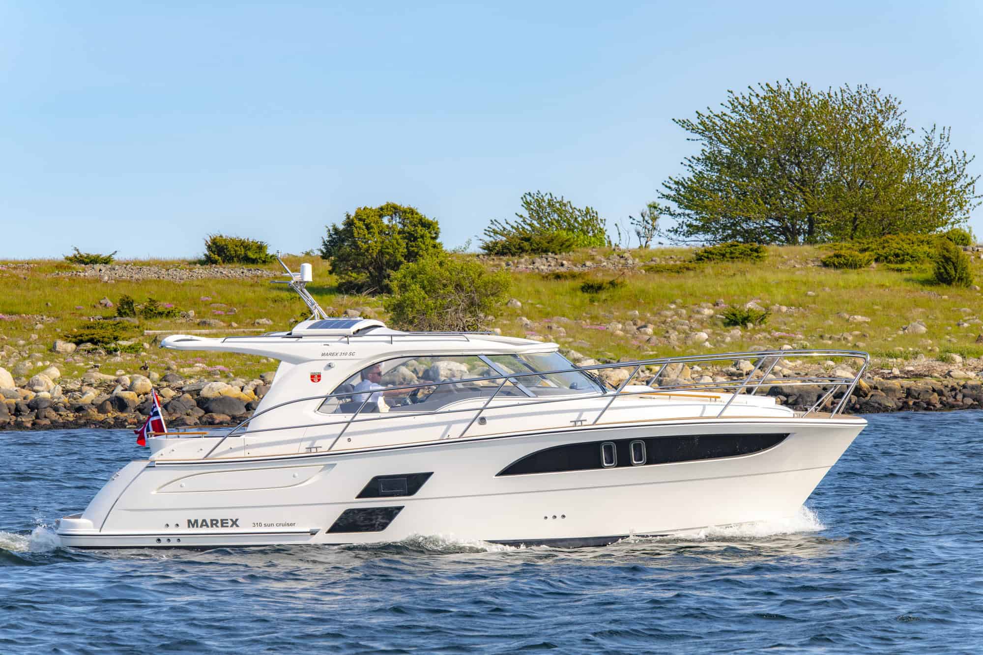 Biały jacht motorowy Marex 310 Sun Cruiser płynący po wodzie w słoneczny dzień, z zielonym brzegiem w tle.