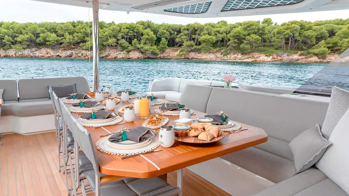 Duży stół jadalny na zewnętrznym pokładzie katamaranu Fountaine Pajot Thíra 80, z widokiem na morze i zalesiony brzeg.