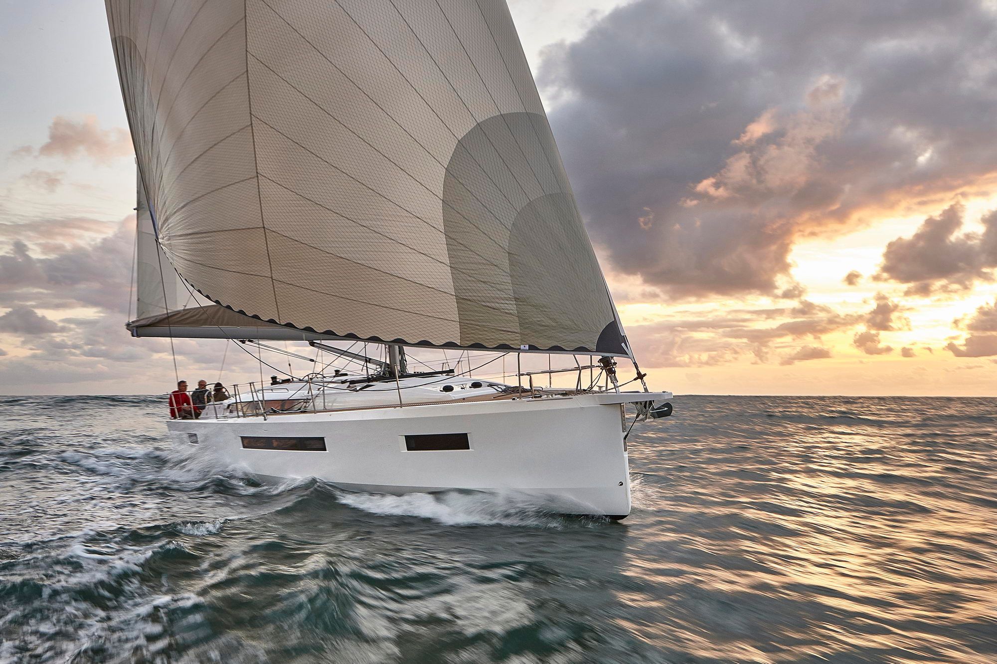 Jacht żaglowy Jeanneau Sun Odyssey 490 płynący po morzu o zachodzie słońca. Widoczny dziób, żagle i załoga na pokładzie.