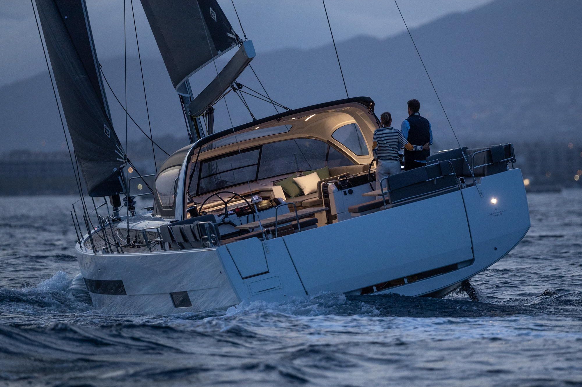 Jacht żaglowy Jeanneau Yachts 55 płynący po morzu o zmierzchu, z dwoma osobami w kokpicie i podniesionymi żaglami.