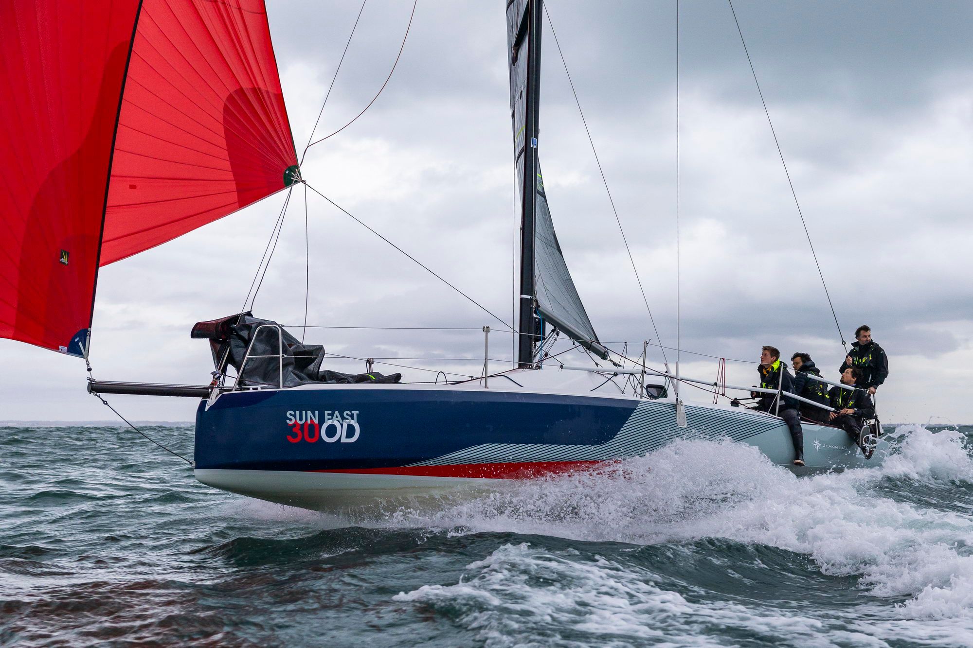 Jeanneau Sun Fast 30 One Design auf bewegtem Wasser mit gesetztem rotem Spinnaker und Crew an der Reling.