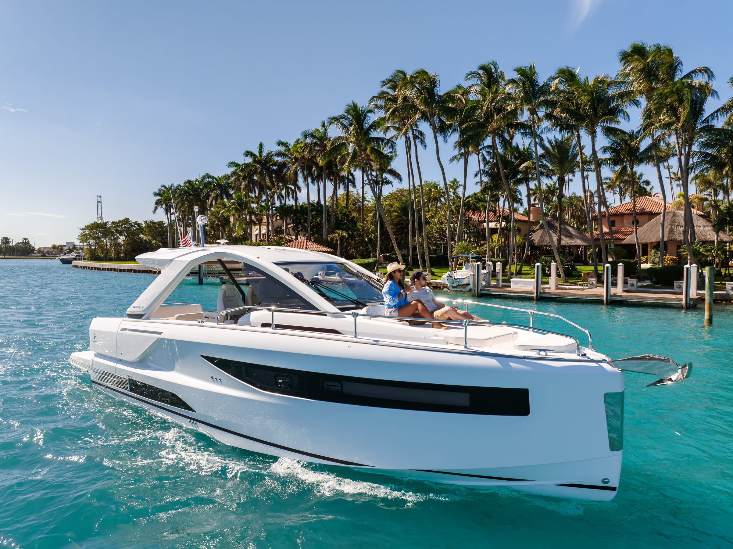 White Jeanneau DB/37 OB motorboat with two people on the bow, cruising on turquoise water on a sunny day.