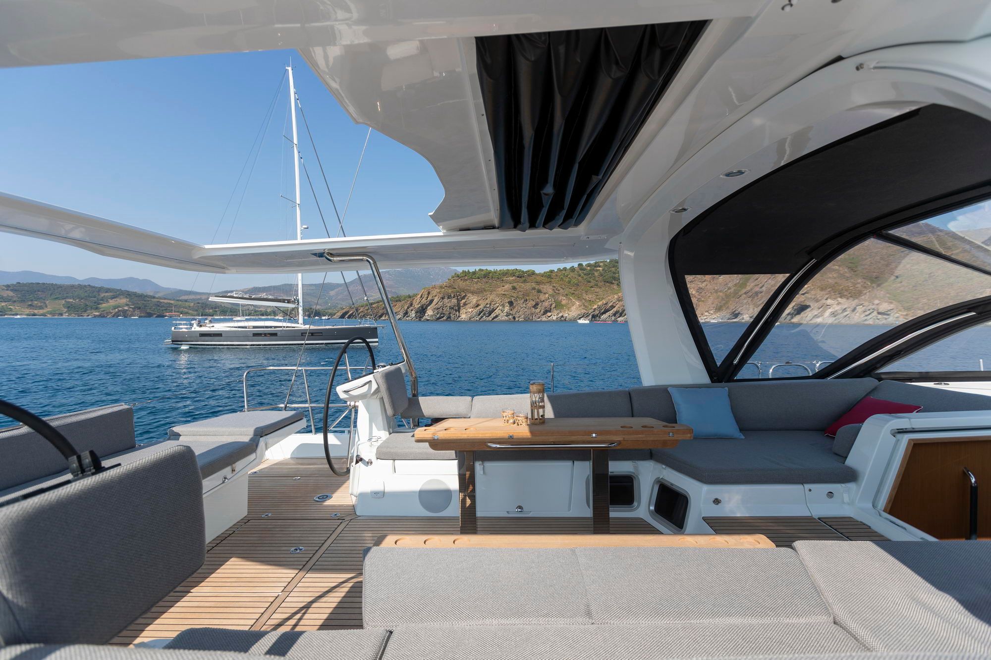 Przestronny kokpit jachtu Jeanneau Yachts 60 z drewnianym stołem, siedziskami i widokiem na morze oraz inną żaglówkę.