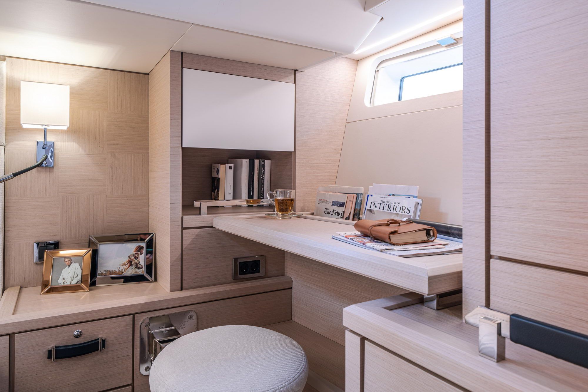 Jasne wnętrze kabiny jachtu Jeanneau Yachts 65 z biurkiem, szafkami, lampą i oknem. Widoczne książki i dekoracje.