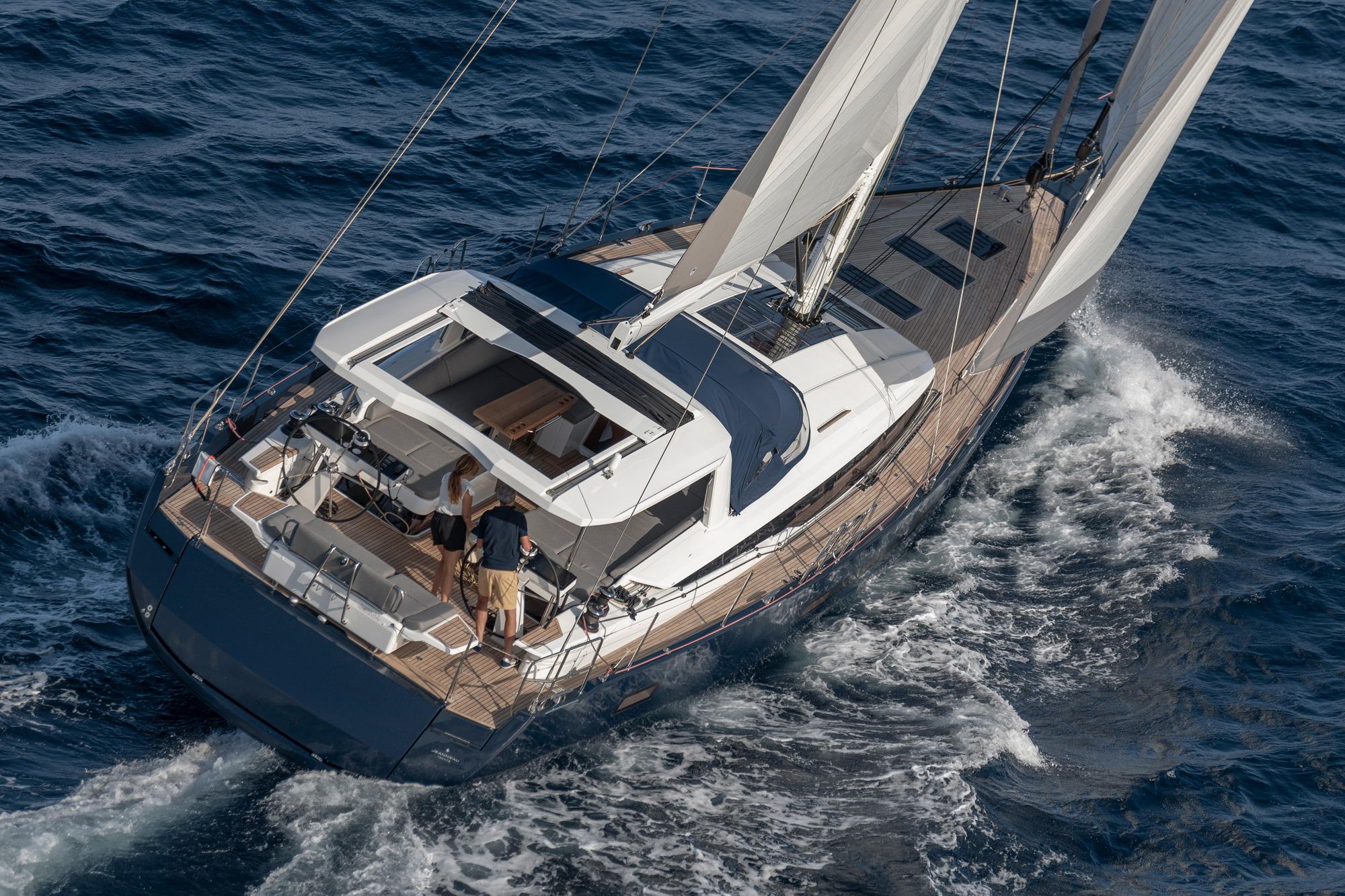 Widok z góry na jacht żaglowy Jeanneau Yachts 65 płynący po morzu. Dwie osoby w kokpicie, żagle rozwinięte.