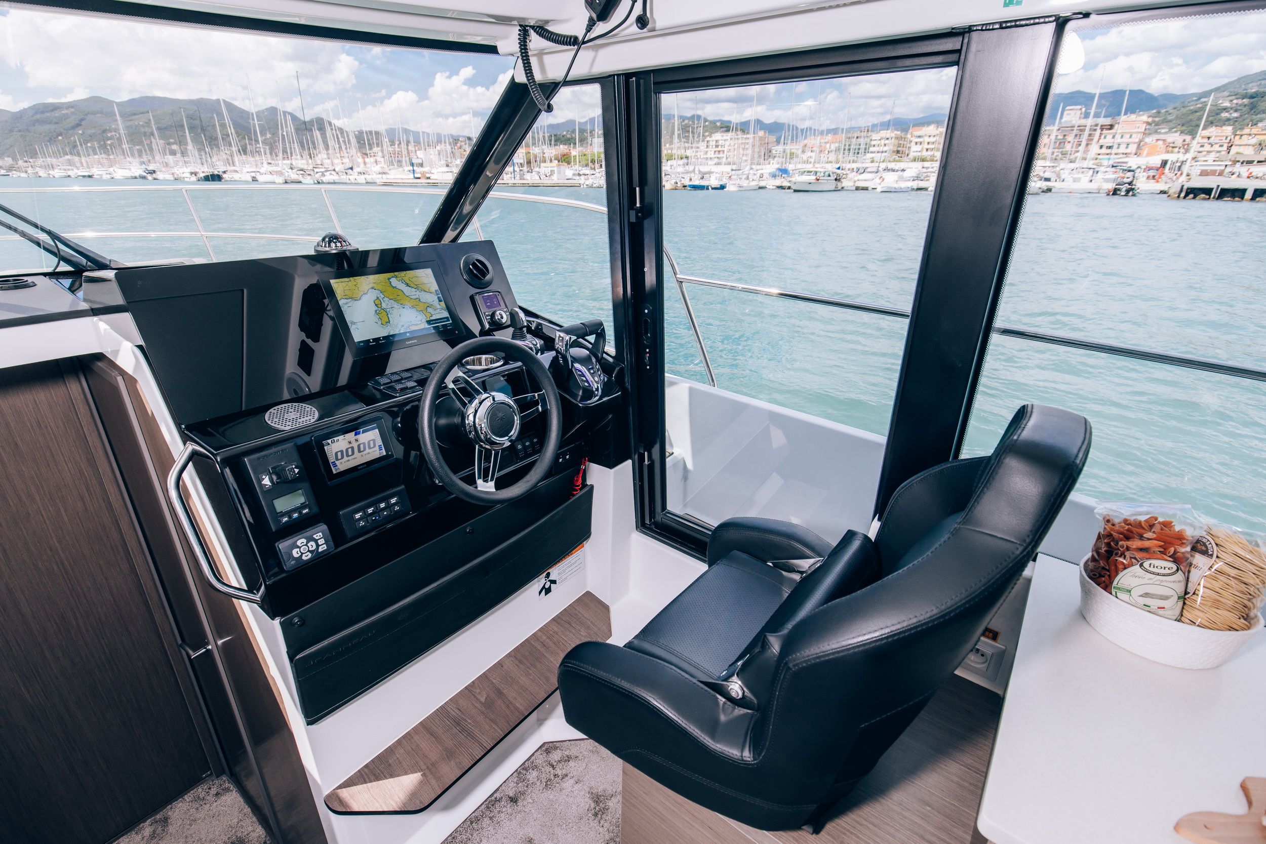 Interior of the Jeanneau Merry Fisher 1095 Fly Serie2, view of the helm station with navigation display and wheel, with a window overlooking the marina.