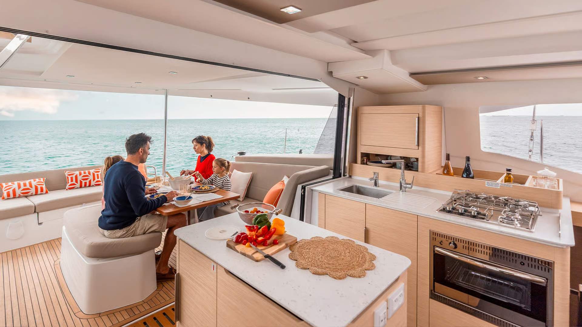 A family eats a meal in the spacious aft cockpit aboard the Fountaine Pajot FP41 catamaran, with sea views. A fully equipped galley is also visible.