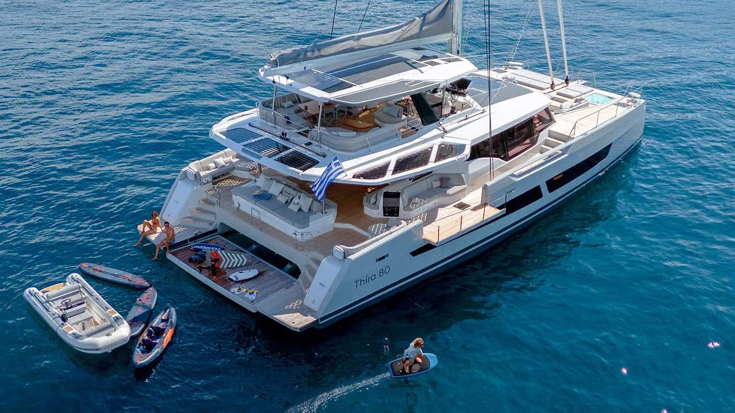 Widok z lotu ptaka na luksusowy katamaran Fountaine Pajot Thíra 80 z otwartymi balkonami bocznymi i ludźmi na rufie.