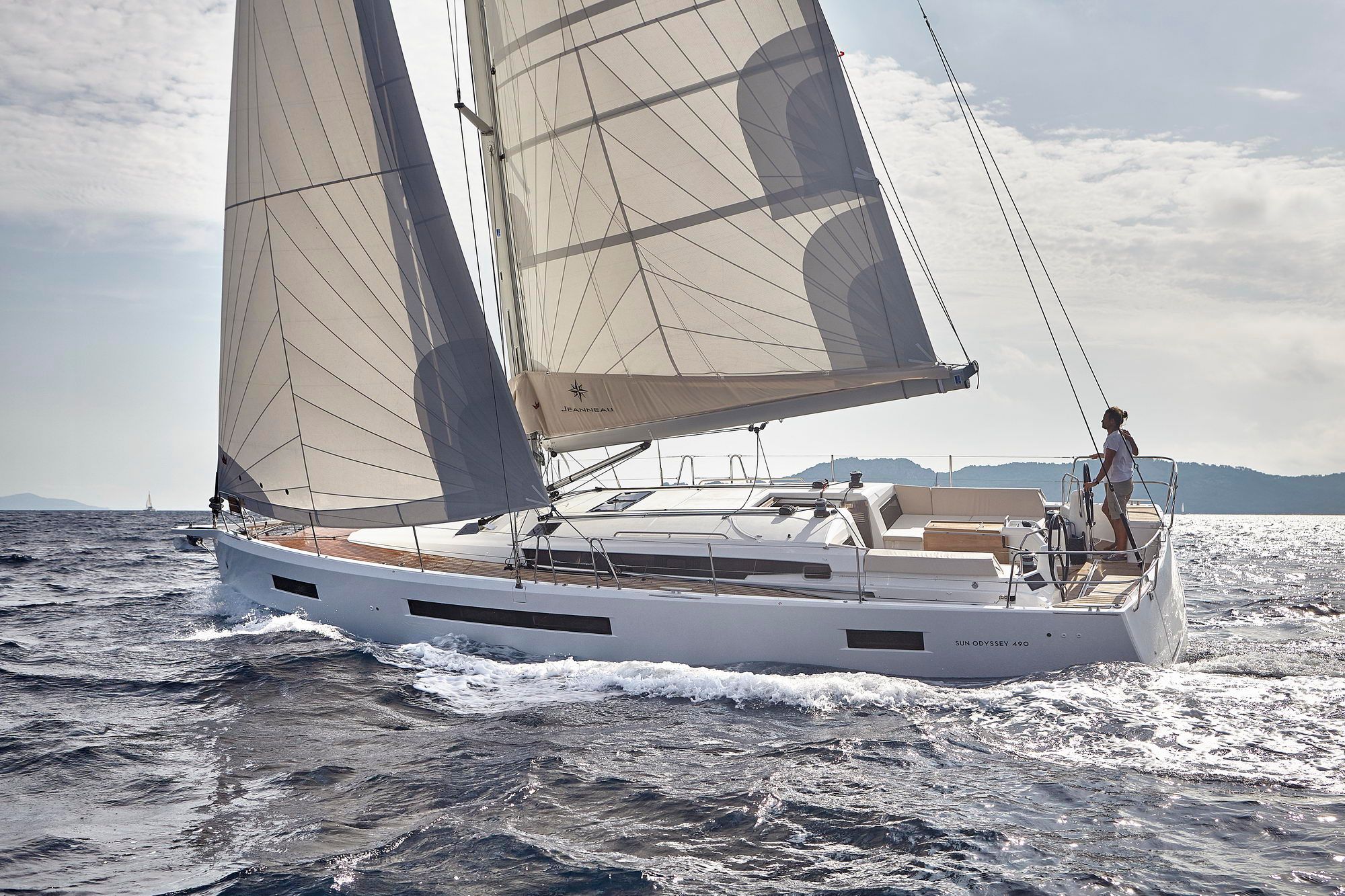 Biała żaglówka Jeanneau Sun Odyssey 490 płynąca po morzu z rozłożonymi żaglami. Sternik stoi przy sterze.