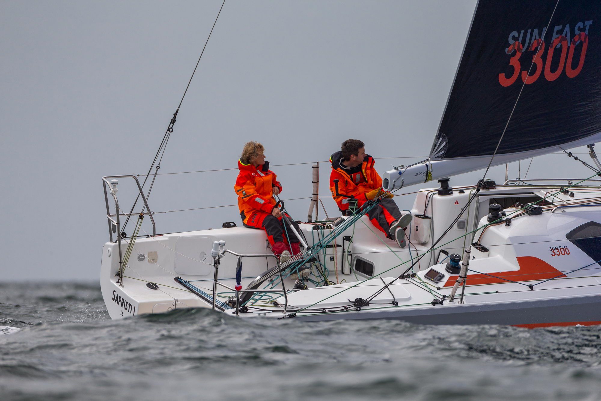 Two sailors in orange jackets on board the Jeanneau Sun Fast 3300 racing yacht while sailing.