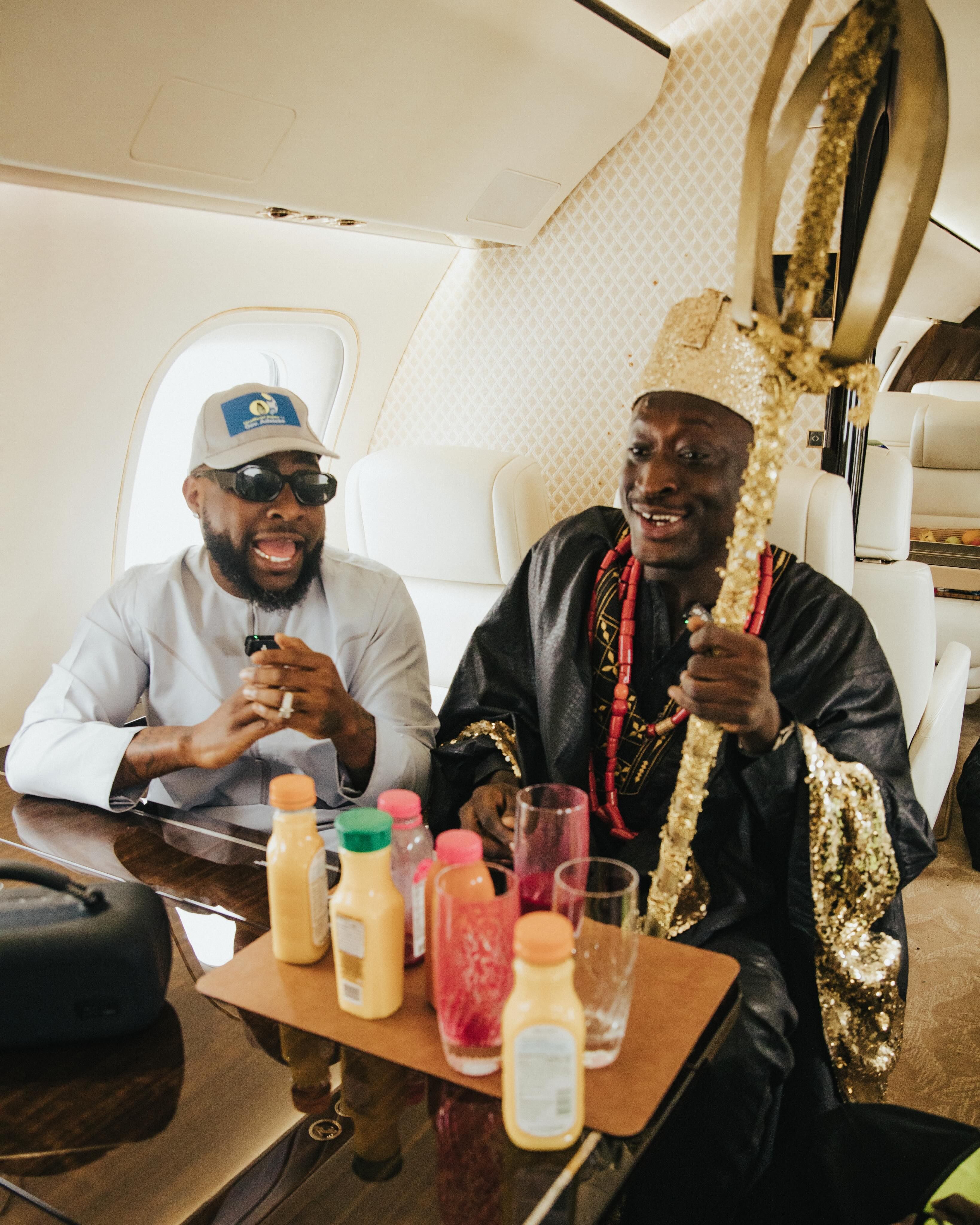 Two men sit inside a private jet, smiling and talking beside a table of bottled drinks and glasses. One wears a white outfit, cap, and sunglasses, while the other is dressed in black traditional attire with red beads and holds an ornate gold staff.