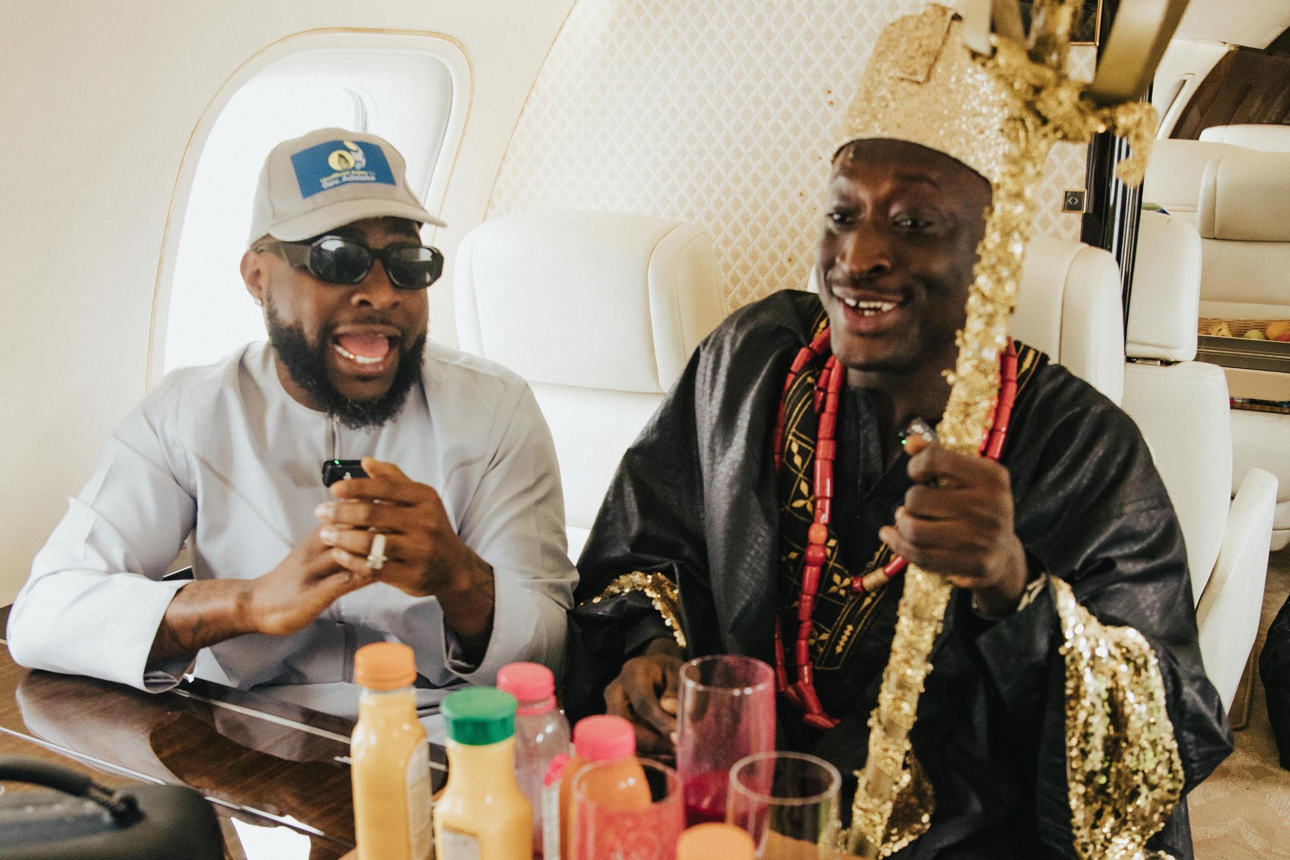 Two men sit inside a private jet, smiling and talking beside a table of bottled drinks and glasses. One wears a white outfit, cap, and sunglasses, while the other is dressed in black traditional attire with red beads and holds an ornate gold staff.