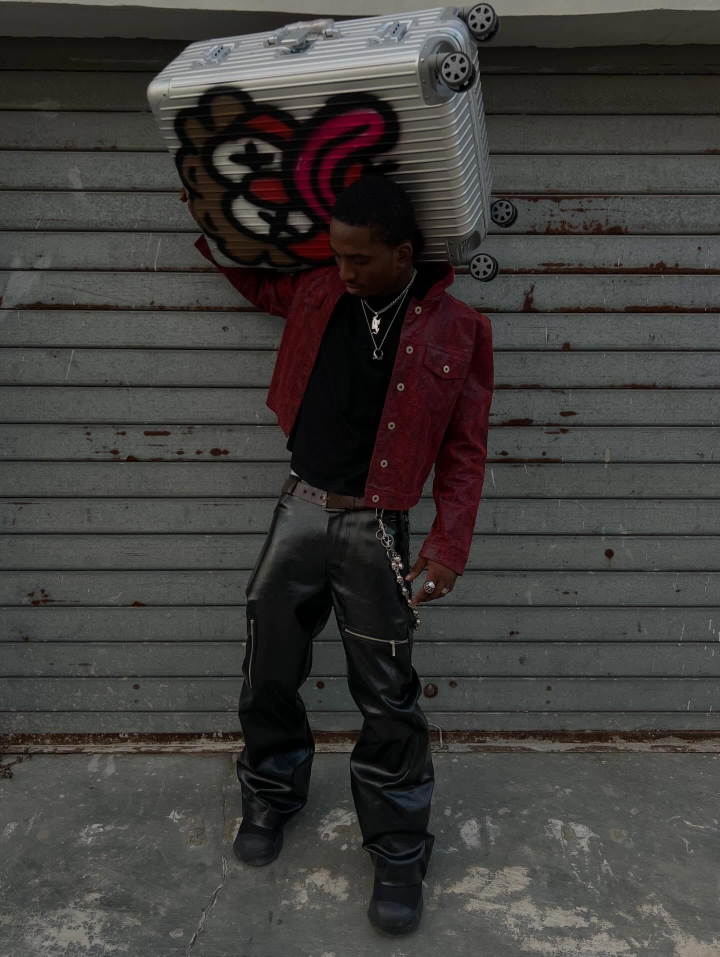 A young man poses in front of a metal shutter, wearing a red jacket, black leather trousers, layered necklaces and dark shoes, while carrying a large silver suitcase covered with colorful graffiti-style artwork on his shoulder.