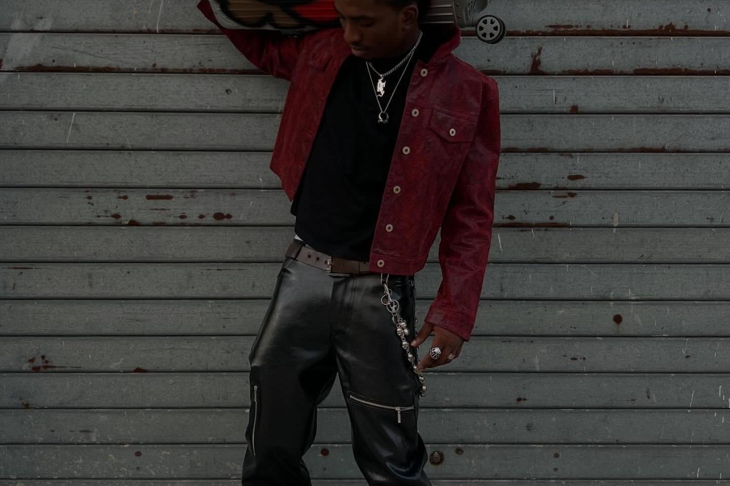 A young man poses in front of a metal shutter, wearing a red jacket, black leather trousers, layered necklaces and dark shoes, while carrying a large silver suitcase covered with colorful graffiti-style artwork on his shoulder.