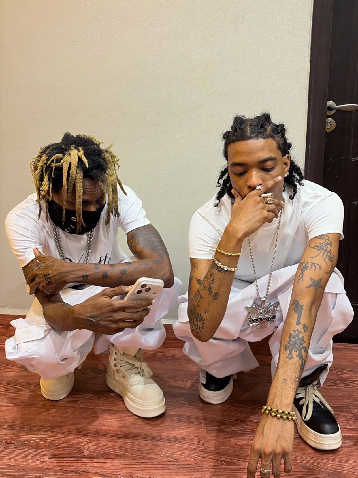 Two tattooed men crouch on a wooden floor in coordinated white outfits. One has blond-tipped dreadlocks, wears a black face mask and looks at a phone, while the other rests his hand over his mouth and wears layered jewelry with black-and-white shoes.
