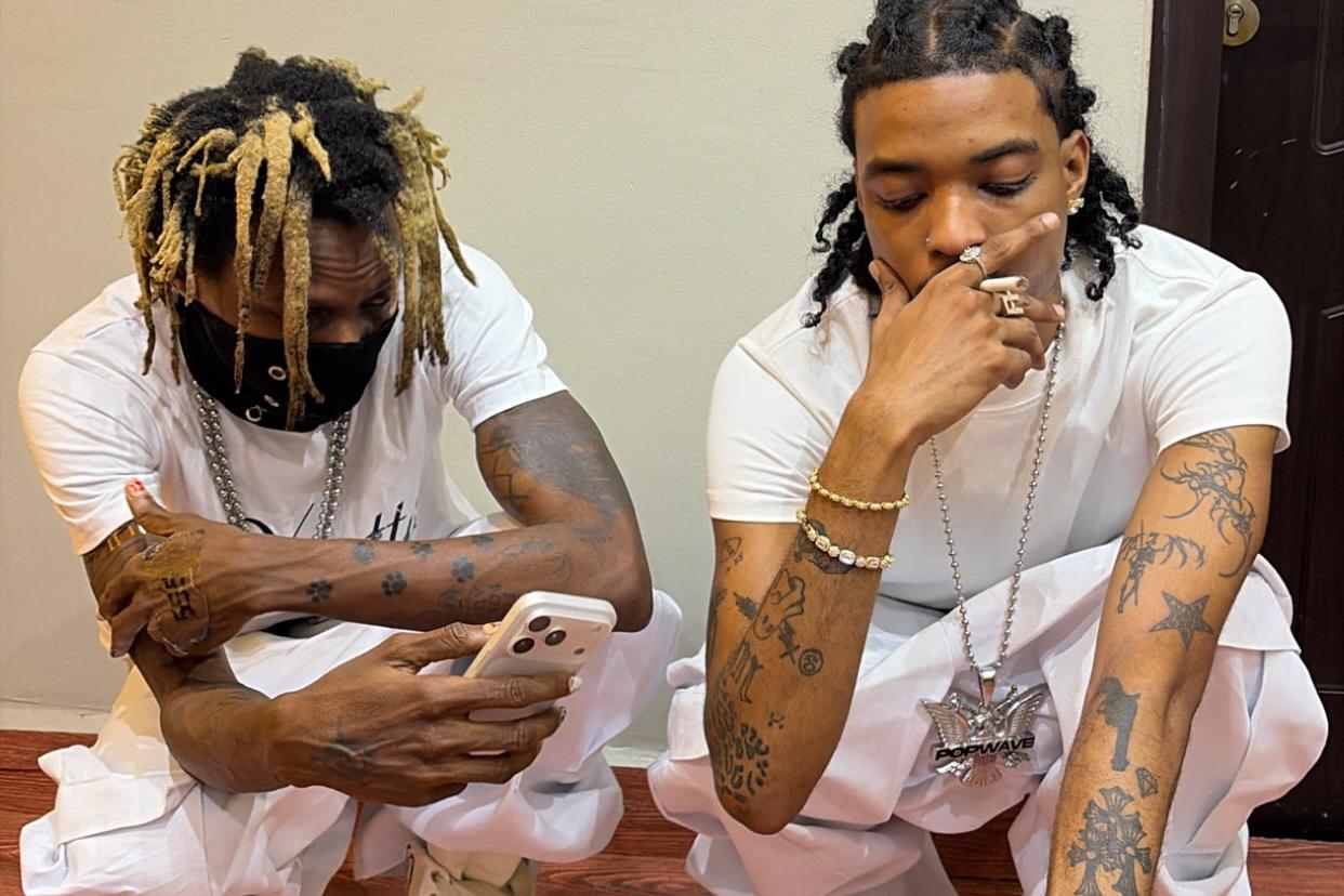 Two tattooed men crouch on a wooden floor in coordinated white outfits. One has blond-tipped dreadlocks, wears a black face mask and looks at a phone, while the other rests his hand over his mouth and wears layered jewelry with black-and-white shoes.