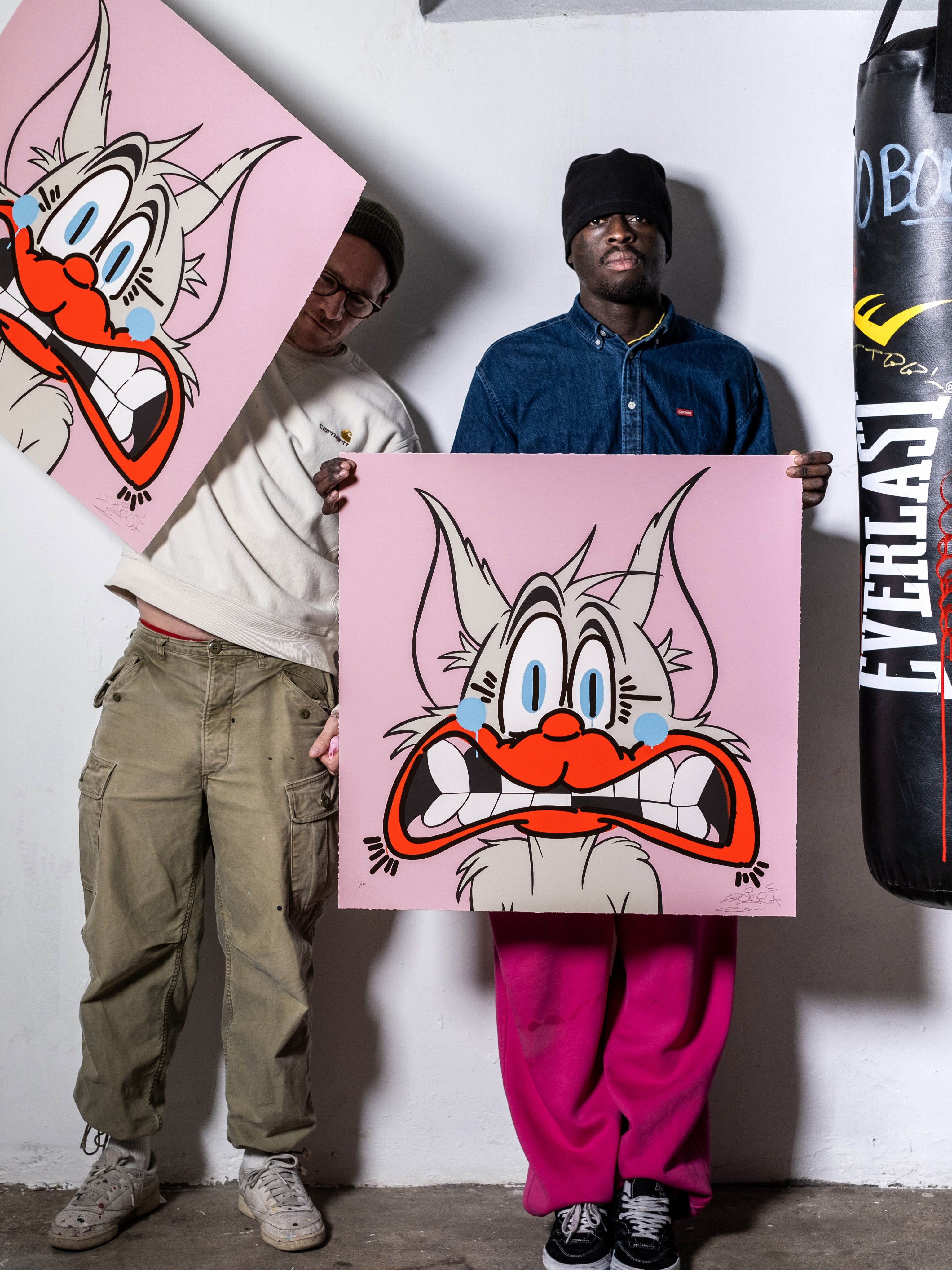 Two men stand in a studio holding large pink artworks featuring an