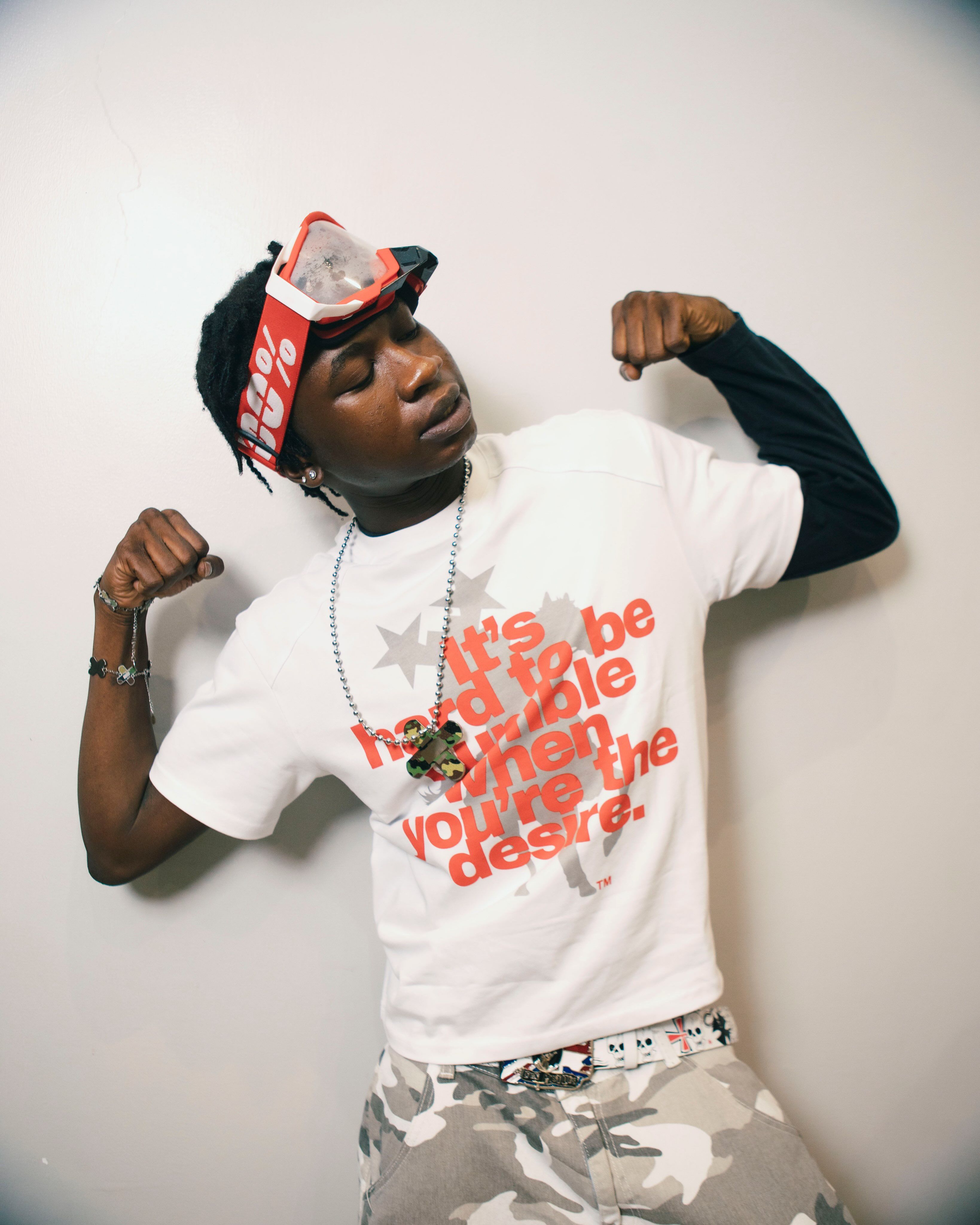ScottyOlorin poses against a plain white wall, flexing both arms while wearing a white graphic T-shirt over a black long-sleeve top, grey camouflage pants, a red visor with goggles, and layered accessories.