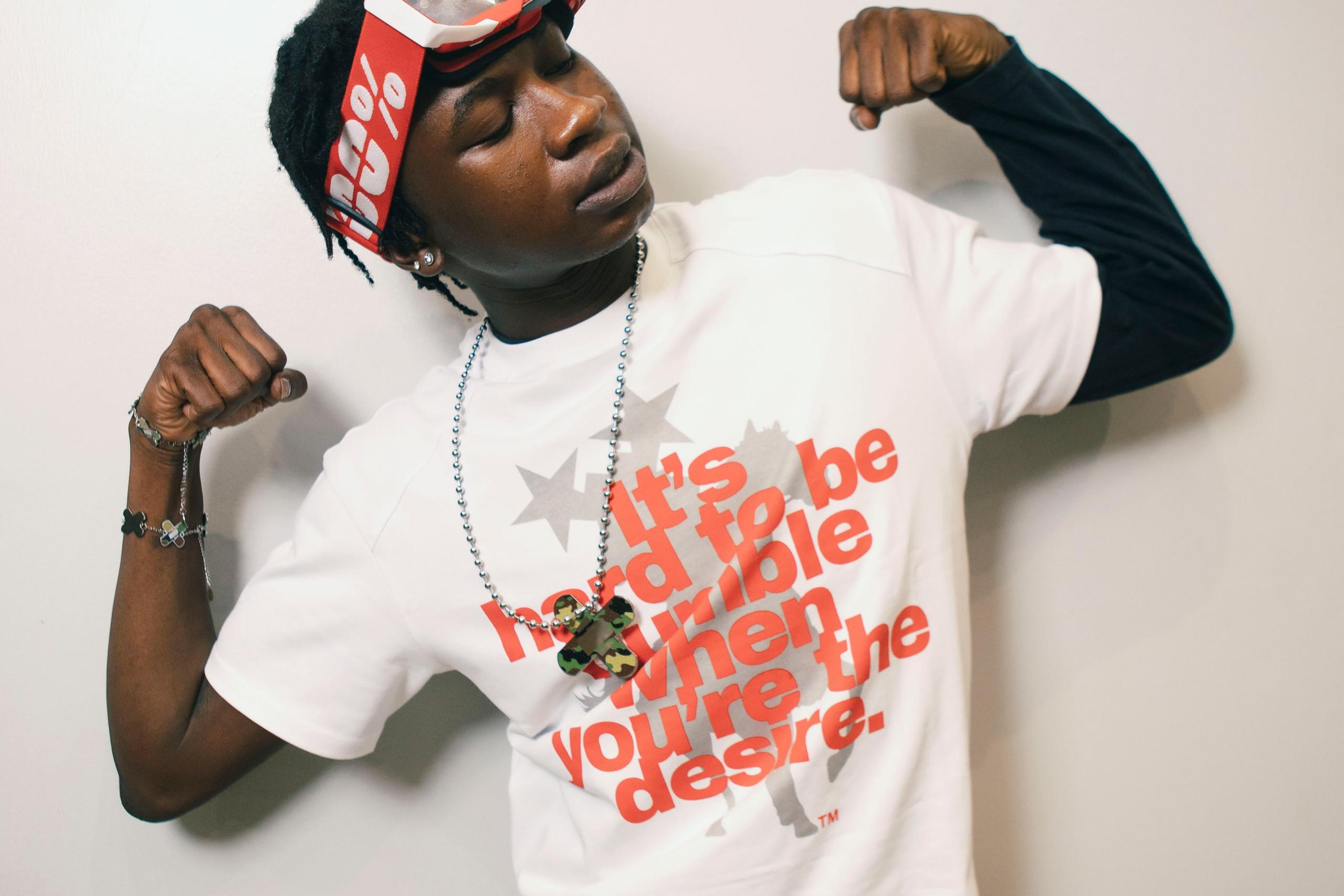 ScottyOlorin poses against a plain white wall, flexing both arms while wearing a white graphic T-shirt over a black long-sleeve top, grey camouflage pants, a red visor with goggles, and layered accessories.